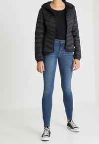 Veste noire matelassée avec capuche, tissu texturé, fermeture éclair à l'avant, associée à un jean skinny bleu foncé et des baskets noires avec des lacets blancs.