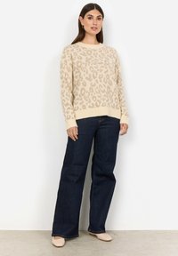 Beige leopardmönstrad tröja med ribbade ärmslut och nederkant, i kombination med mörkblå utsvängda jeans och ljusa ballerina-skor mot en enfärgad bakgrund.