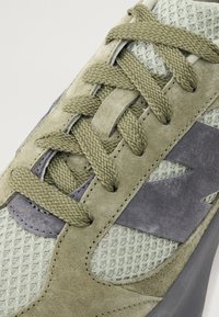 Gros plan d'une basket verte en daim et mesh avec des lacets texturés et un détail gris foncé sur le côté.