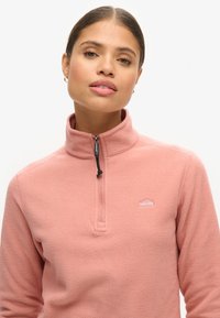 Pullover in pile di colore rosa chiaro, con colletto a mezza zip, logo ricamato sul petto e una consistenza morbida con una vestibilità comoda.