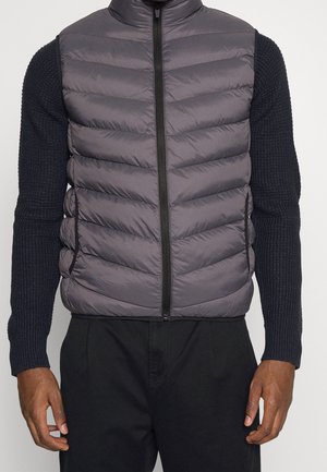 Homme portant une doudoune matelassée gris foncé avec une fermeture éclair noire à l'avant, par-dessus un pull en maille noir à manches longues et un pantalon noir.