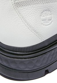 Vita lädersko med texturerad yta, med en svart gummisula. Anmärkningsvärda detaljer inkluderar präglad branding och kontrasterande sömmar.