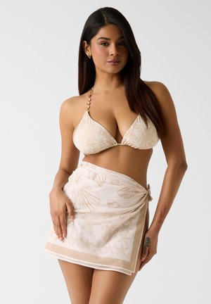 Kobieta o długich ciemnych włosach, ubrana w beżowy top bikini z wzorem oraz pasujący sarong z motywami muszelek i rozgwiazd.