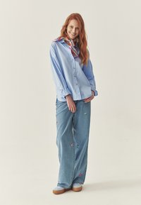 Lichtblauwe gestreepte shirt, comfortabele pasvorm, rode knopen. Wijde blauwe jeans met bloemborduursels, gecombineerd met blauwe schoenen met tan kleurige zolen.