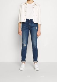 Tmavě modré džínové kalhoty s úzkým střihem, zdobené roztrhanými detaily. V kombinaci s bílým tričkem a lehkou zkrácenou bundou. Bílé tenisky.