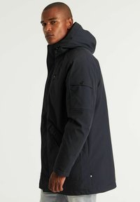 Tamna vodootporna jakna s kapuljačom, s zatvaranjem na patentni zatvarač, bočnim džepovima i teksturiranim materijalom. Podesive manžete i detalj s logotipom na rukavu.