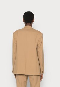 Béžový blazer s přiléhavým střihem, strukturovanými rameny a detailem zadního ventilu. Vyroben z hladké tkaniny, s dlouhými rukávy a moderním střihem.