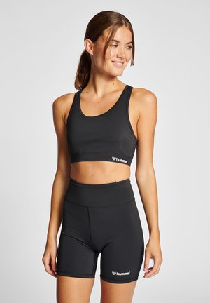 Kobieta stoi, ubrana w czarny sportowy biustonosz i wysokie krótkie spodenki rowerowe z logo "hummel" na obu, uśmiechnięta i patrzy w prawo.