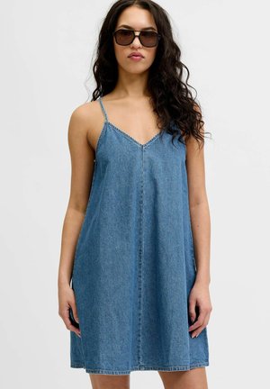 Femme aux longs cheveux bouclés portant de grandes lunettes de soleil noires et une robe en jean bleu sans manches avec de fines bretelles.