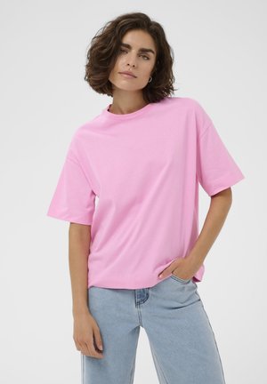 Fiatal nő rövid, göndör hajjal, laza rózsaszín pólót és világoskék farmert visel, egyik kezét zsebre téve pózol fehér háttér előtt.