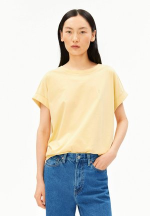 Femme aux longs cheveux noirs portant un t-shirt jaune clair à manches courtes rentré dans un jean taille haute bleu, une main dans la poche.