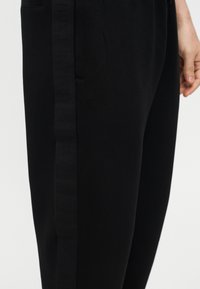 Κοντινό πλάνο μαύρου παντελονιού με διακριτική λεπτομέρεια πλαϊνής ρίγας και μερική όψη του χεριού ενός ατόμου δίπλα στο παντελόνι.