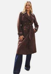 Rudas suede trench paltas su dvigubos rankovėmis, surištas diržas, antpečiai ir įpjovimu dekoruotas apykaklė. Dėvimas ant tamsaus džinsinio audinio kelnių.