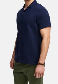 Uomo che indossa una camicia blu navy a maniche corte e pantaloni verde oliva, con un braccialetto di perline sul polso destro.