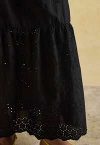 Czarna bawełniana sukienka z haftowanym kwiatowym wzorem i falowanym dołem, z warstwowym fasonem. Obuwie to żółte sandały.