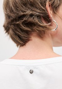 Λευκό μπλουζάκι με στρογγυλή λαιμόκοψη και κοντά μανίκια, διακοσμημένο με μια μικρή ασημένια κουμπί στο πίσω μέρος του λαιμού. Λαχταριστός βαμβακερός ύφασμα.