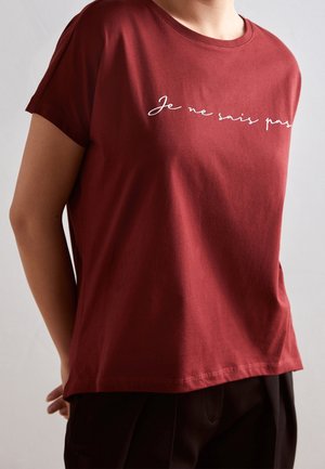 Person trägt ein locker sitzendes dunkelrotes T-Shirt mit weißem Schriftzug „Je ne sais pas“ über der Brust, kombiniert mit schwarzen Hosen.