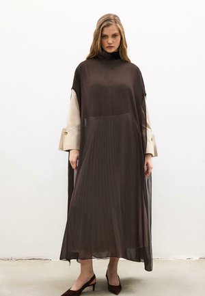 Femme portant une longue robe noire transparente par-dessus une chemise beige, debout devant un fond blanc uni, regardant droit devant avec une expression neutre.