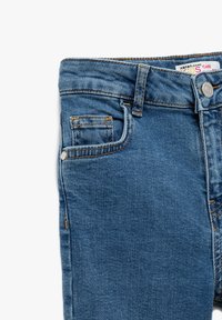 Flareshousut keskisinisestä denimistä. Viiden taskun malli, jossa on oranssia tikkausta ja metallinen nappi. Sileä pinta, hieman haalistunut viimeistely.