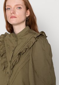 Camisa verde-azeitona com detalhes em renda, colarinho alto e fecho com botões. Apresenta um tecido texturizado e costura decorativa ao longo das costuras.