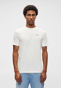 Balta kokvilnas T-kreklā ar īsām piedurknēm, apaļu dekoltē un mazs melns logo uz kreisās krūtis. Vienkāršs un klasiskas dizains.