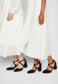 Czarne zamszowe buty na słupku z krzyżowymi paskami i spiczastymi czubkami, noszone z płynącymi białymi sukienkami. Gładka i matowa faktura.