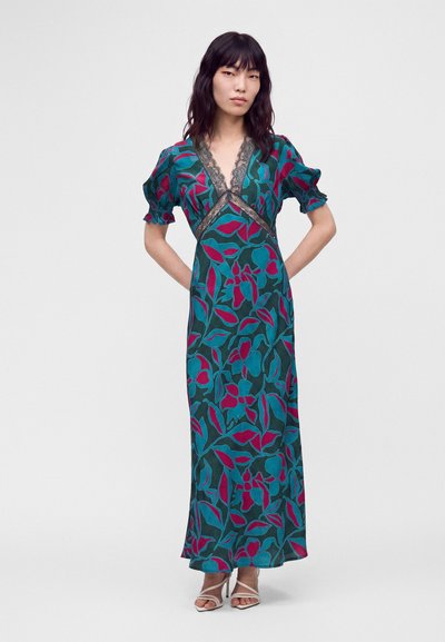 Femme portant une robe maxi fleurie bleu sarcelle et magenta avec un bord en dentelle au niveau du décolleté et des manches courtes bouffantes, debout les mains dans le dos.