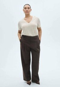 T-shirt beige à col en V associé à un pantalon large marron taille haute avec des plis devant. Le mannequin a un look de maquillage naturel et porte des boucles d'oreilles créoles.