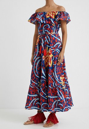 Femme portant une longue robe à motifs bleus et rouges, épaules dénudées, ceinturée de pompons, assortie à des sandales plates rouges à franges.