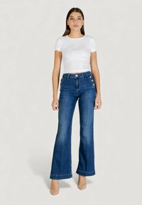 Top bianco a maniche corte con texture e logo "Guess" in argento, abbinato a jeans svasati blu scuro con bottoni decorativi in vita.