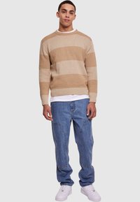 Pulover bej cu dungi, cu mansete și tiv cu nervuri, purtat peste un tricou alb; asortat cu blugi denim de un albastru deschis și adidași albi.