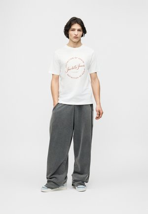 Junger Mann steht mit den Händen in den Taschen, trägt ein weißes Jack & Jones T-Shirt, weite graue Hosen und hellfarbene Turnschuhe vor einem schlichten Hintergrund.