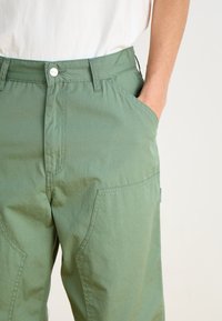 Pantalons en coton vert avec une coupe droite, dotés de poches latérales, d'une fermeture à bouton en métal et de détails de couture visibles.