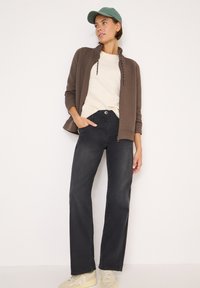 Giacca marrone con zip sopra un maglione beige, abbinata a jeans neri a gamba dritta e sneakers beige. Il modello indossa anche un cappellino verde.
