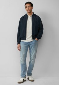 Marine bomberjack, beige poloshirt, lichtblauwe jeans en witte sneakers met zwarte accenten. Gladde stof en een relaxte pasvorm.