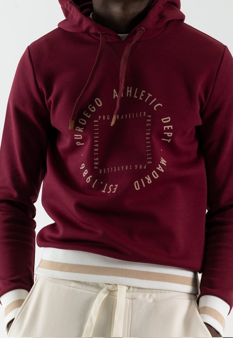 Bordowa bluza z kapturem z okrągłym beżowym logo, z sznurkami ściągającymi i ściągaczami na rękawach. Spodnie są kremowe z beżowym paskiem w paski w talii.