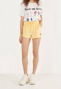 Gula dragkedjeshorts i mjukt tyg med en broderad fjäril. Kombineras med en vit grafisk T-shirt med färgglada juvelillustrationer.
