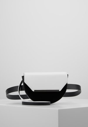 Sort og hvid halvmåneformet crossbody-taske med sort rem, placeret på en hvid sokkel mod en ensfarvet lys grå baggrund.