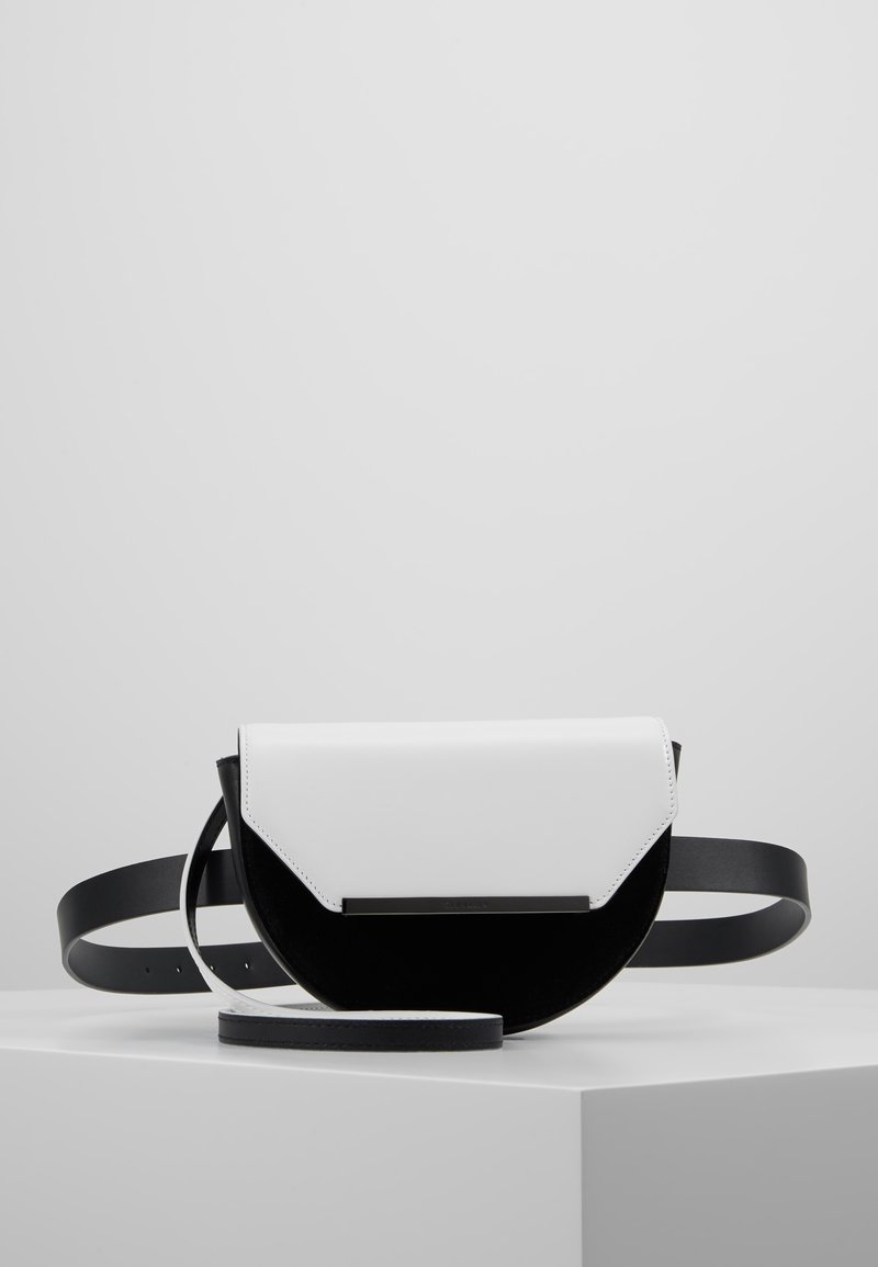 Sac bandoulière en forme de demi-lune noir et blanc avec une sangle noire, placé sur un socle blanc sur un fond gris clair uni.