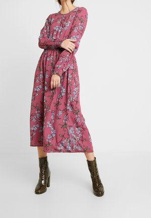 Femme portant une robe midi fleurie violette à manches longues, associée à des bottines marron foncé à talons hauts et texture crocodile, debout, les bras croisés.