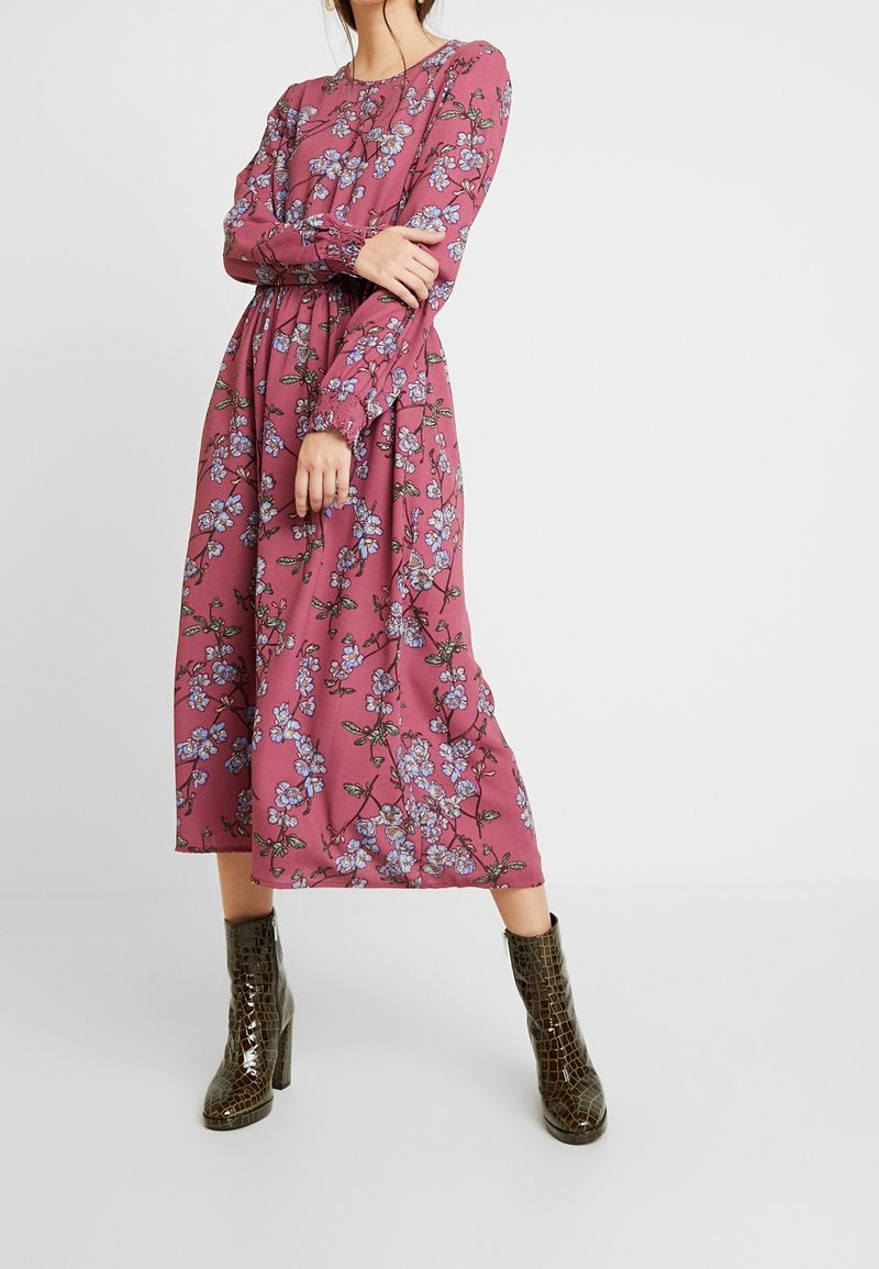 Femme portant une robe midi fleurie violette à manches longues, associée à des bottines marron foncé à talons hauts et texture crocodile, debout, les bras croisés.