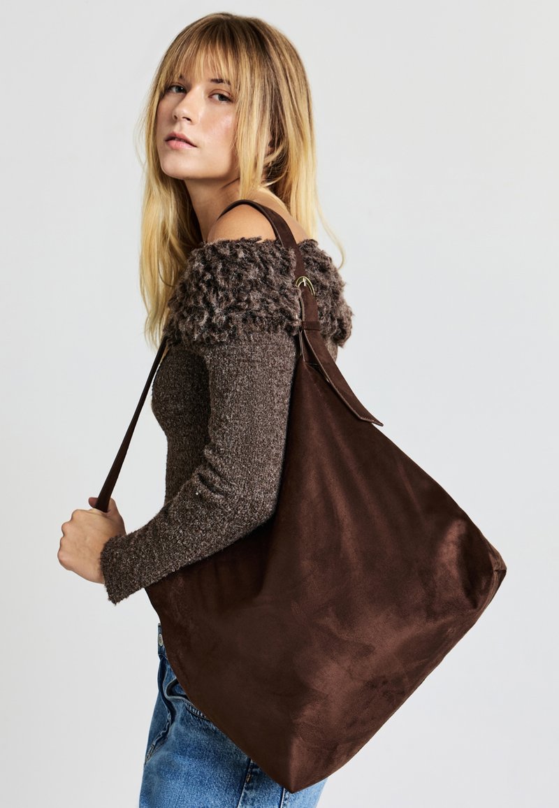 Braune Wildleder-Tasche mit glatter Textur und breiten Schulterriemen. Dazu wird ein weicher, flauschiger brauner Strickpullover schulterfrei getragen.