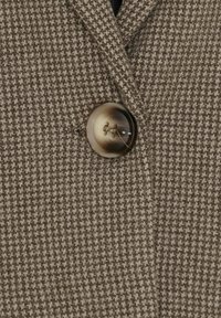 Brun og beige hundetandsmønstret stof i nærbillede med en stor, rund, firehuls knap syet i midten.