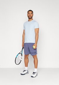 Camiseta de manga corta azul claro, pantalones cortos de deporte morados con el logo "BORG", sosteniendo una pelota de tenis y una raqueta, vistiendo zapatillas blancas y azul marino.