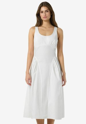 Vrouw met lang bruin haar die een witte mouwloze midi-jurk draagt met een aansluitend lijfje en geplooide zijzakken, staand tegen een effen achtergrond.