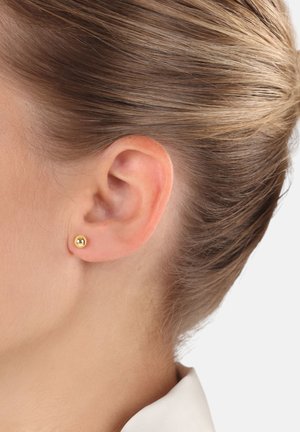 Primer plano de la oreja de una mujer con un pequeño pendiente de bola de oro, cabello castaño claro recogido hacia atrás, vistiendo una camisa con cuello blanca.