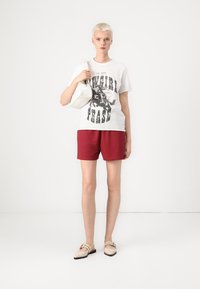 Witte katoenen t-shirt met zwarte grafische tekst, gecombineerd met rode shorts. Model draagt een witte handtas en heeft beige sandalen aan.
