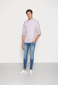 Camicia button-up viola chiaro, indossata sopra una t-shirt bianca. Jeans blu attillati e scarpe slip-on bianche. Tessuto liscio, design casual, dettagli minimi.