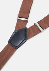Porte-suspente en tissu marron avec une finition texturée, présentants des clips en métal argenté et un accent en cuir noir au point de connexion.