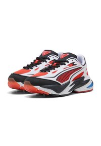 Un paio di scarpe da ginnastica bianche, nere e rosse con tomaia in mesh, lacci neri e dettagli blu sulla suola in gomma bianca, mostrate da un angolo leggermente inclinato.